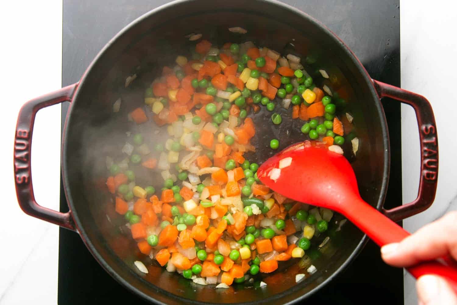 garlic and mixed vegetables added to a pan of sauteed onions, stirred with a red spatula spoon.