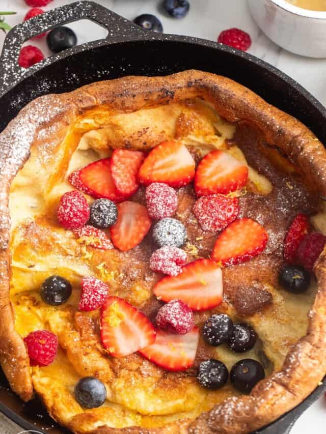 a cast iron skillet holding a german dutch baby pancake topped with powdered sugar, raspberries, blueberries, and sliced strawberries.