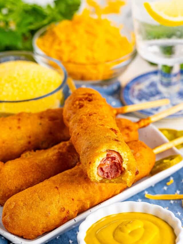 a square plate of four handmade corn dogs with cheddar cheese and jalapenos in the batter. In front of the plate is a small dish of mustard.