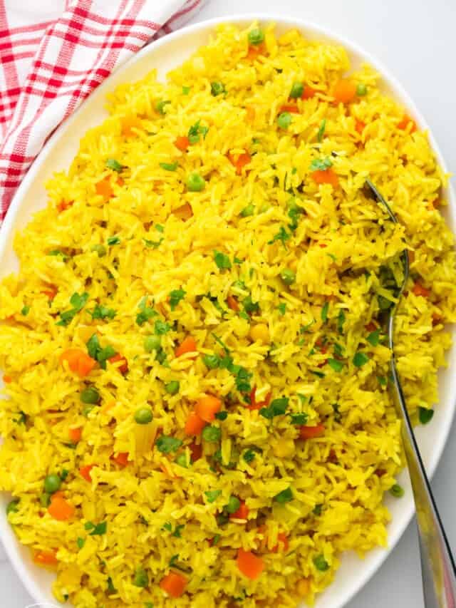 a large oval platter holding tumeric yellow rice with mixed vegetables, garnished with fresh herbs. A serving spoon is in the rice and a red and white plaid towel is above the platter.