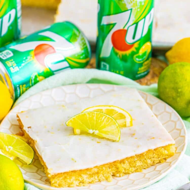 a square slice of 7 up sheet cake with icing, topped with lemon wedges. in the background are cans of 7-up.