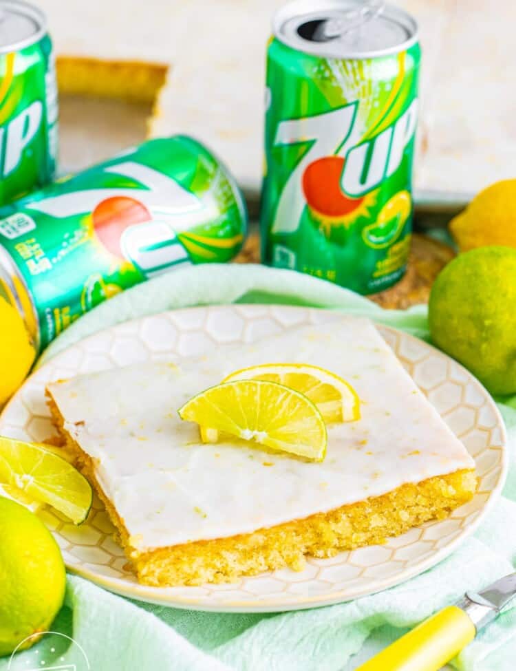a square slice of 7 up sheet cake with icing, topped with lemon wedges. in the background are cans of 7-up.