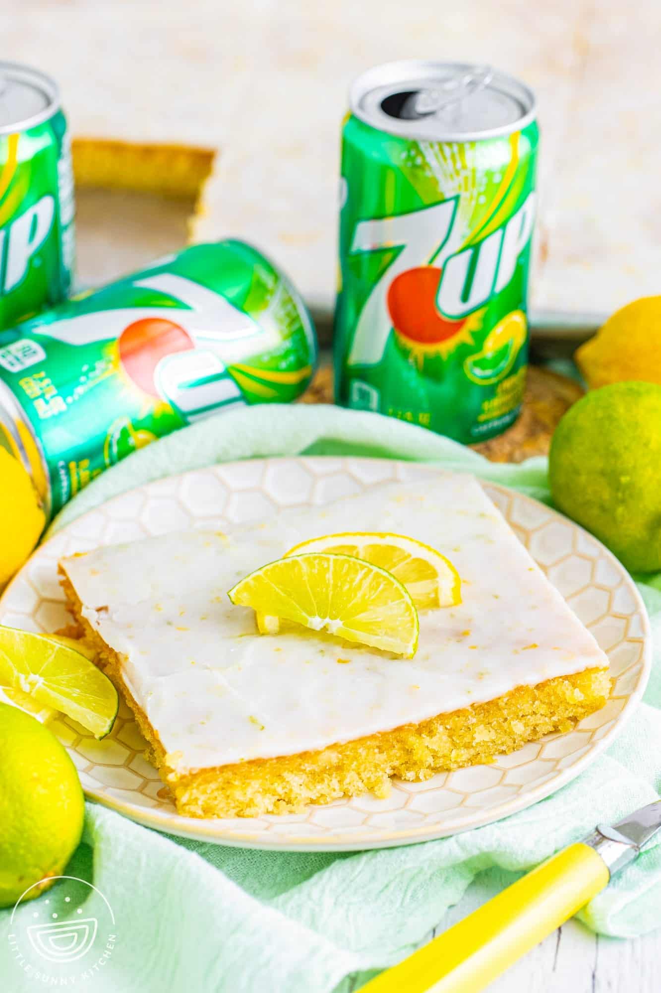a square slice of 7 up sheet cake with icing, topped with lemon wedges. in the background are cans of 7-up. 