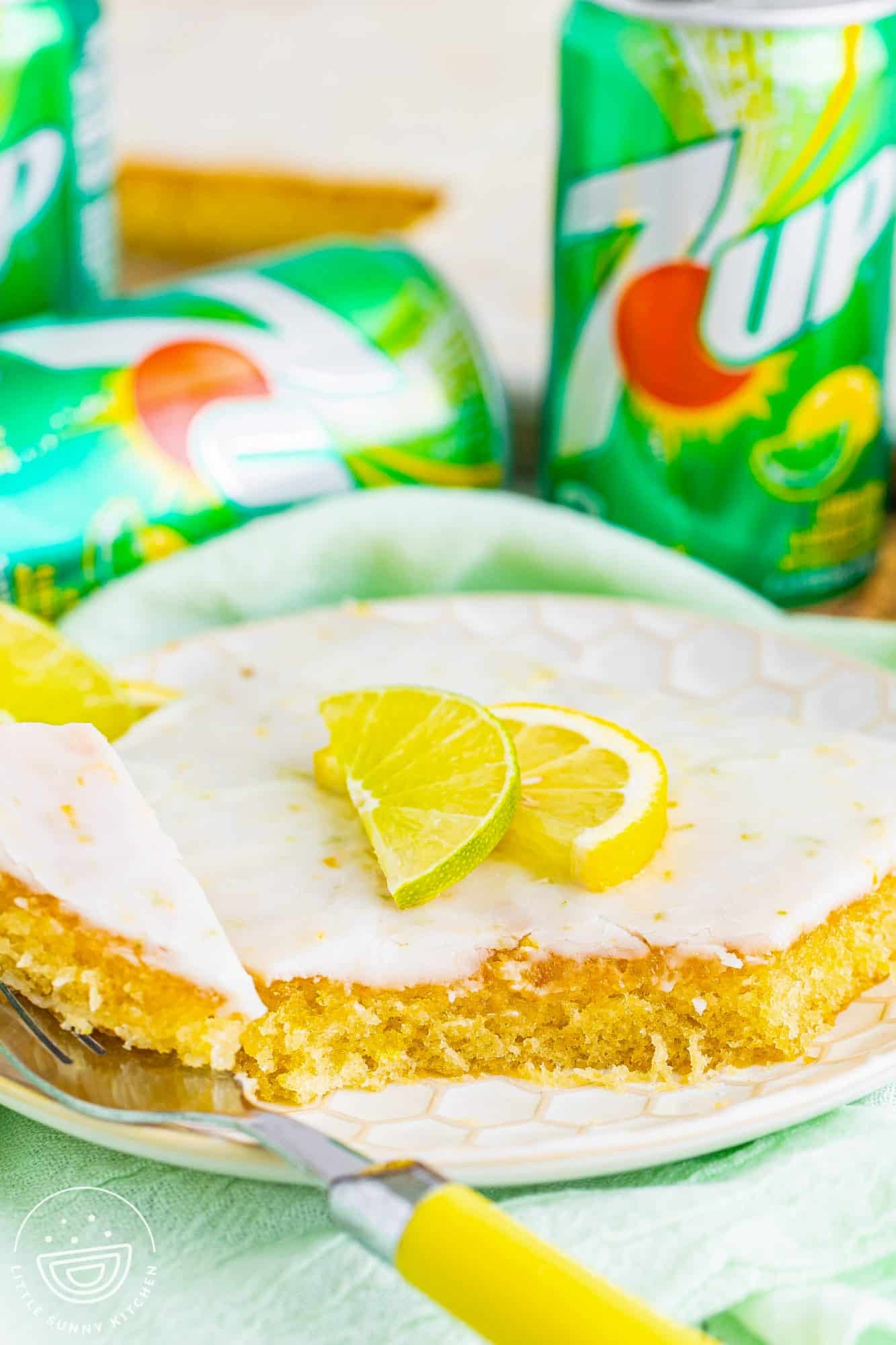 a slice of 7 up sheet cake on a plate. a fork is eating a bite. in the background are cans of pop. 