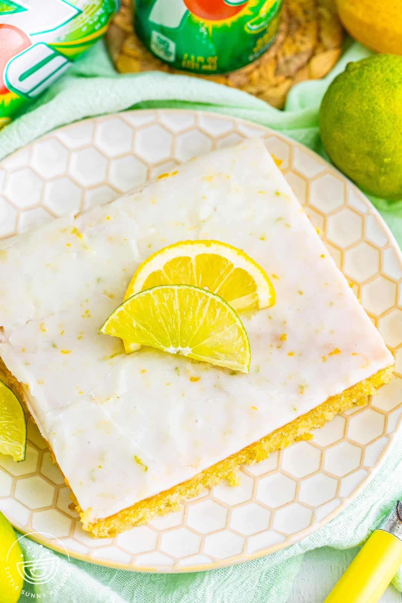 top down view of a square slice of 7up sheet cake on a textured plate. Lemon and lime wedges are added as garnish. 