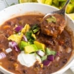 a white bowl of thick soup made with black beans. It's topped with fresh veggies, chips, and sour cream. A spoon is picking up a bite.