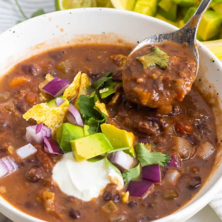 a white bowl of thick soup made with black beans. It's topped with fresh veggies, chips, and sour cream. A spoon is picking up a bite.
