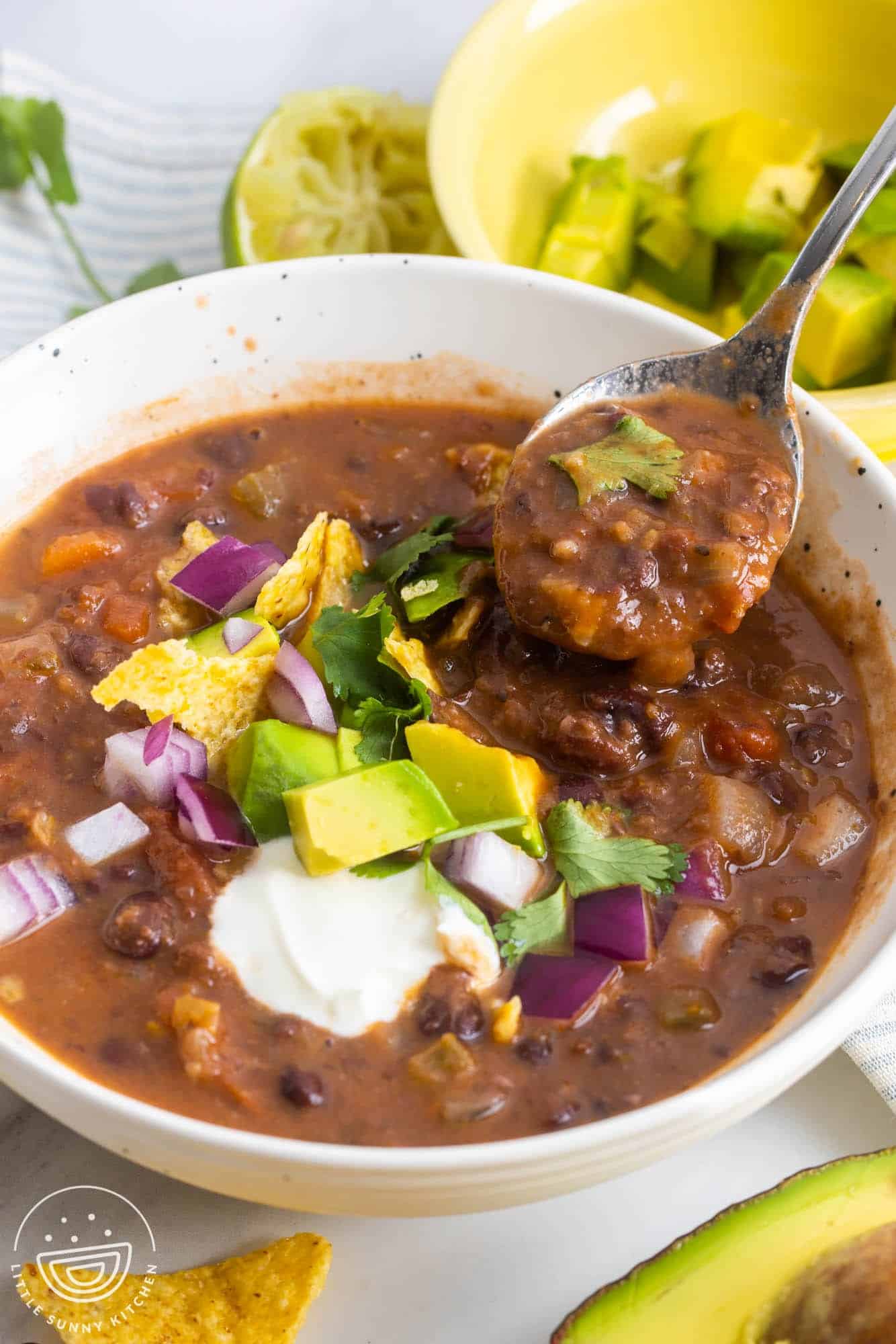 a white bowl of thick soup made with black beans. It's topped with fresh veggies, chips, and sour cream. A spoon is picking up a bite.