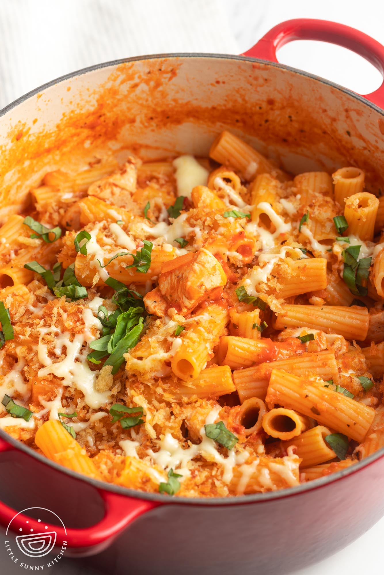 a large dutch oven filled with rigatoni and chicken in a tomato sauce, topped with cheese and breadcrumbs