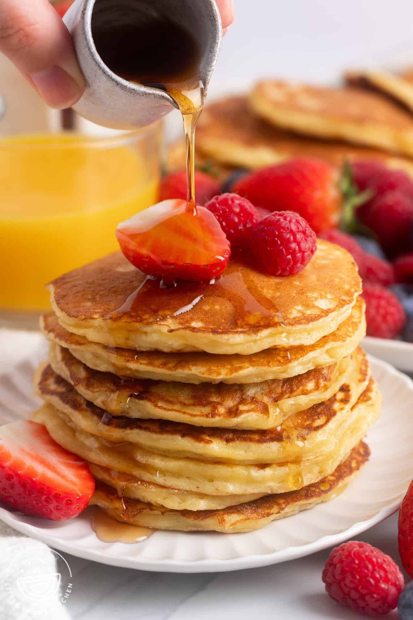 A stack of pancakes with cottage cheese, topped with fresh berries. A hand is pouring maple syrup over the stack. A glass of orange juice is in the background.