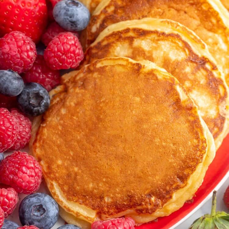 a row of cottage cheese pancakes on a red plate with fresh blueberries and raspberries on the side.