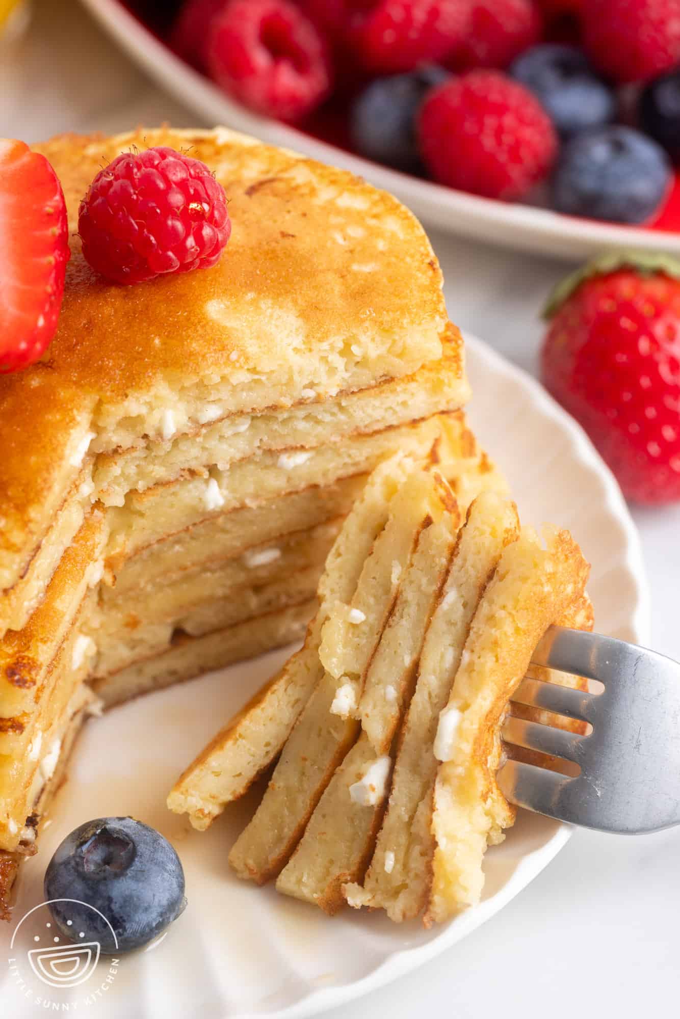 A stack of seven cottage cheese pancakes. The stack has been cut and a fork is holding a bite. On top of the stack is fresh berries and syrup.