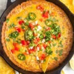 a cast iron skillet filled with hot bean dip topped with cheese, sliced jalapenos, tomatoes, onions, cilantro. Around the pan are tortilla chips.