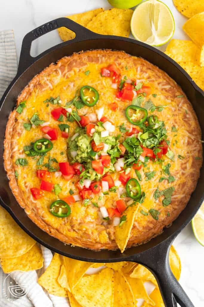 a cast iron skillet filled with hot bean dip topped with cheese, sliced jalapenos, tomatoes, onions, cilantro. Around the pan are tortilla chips.