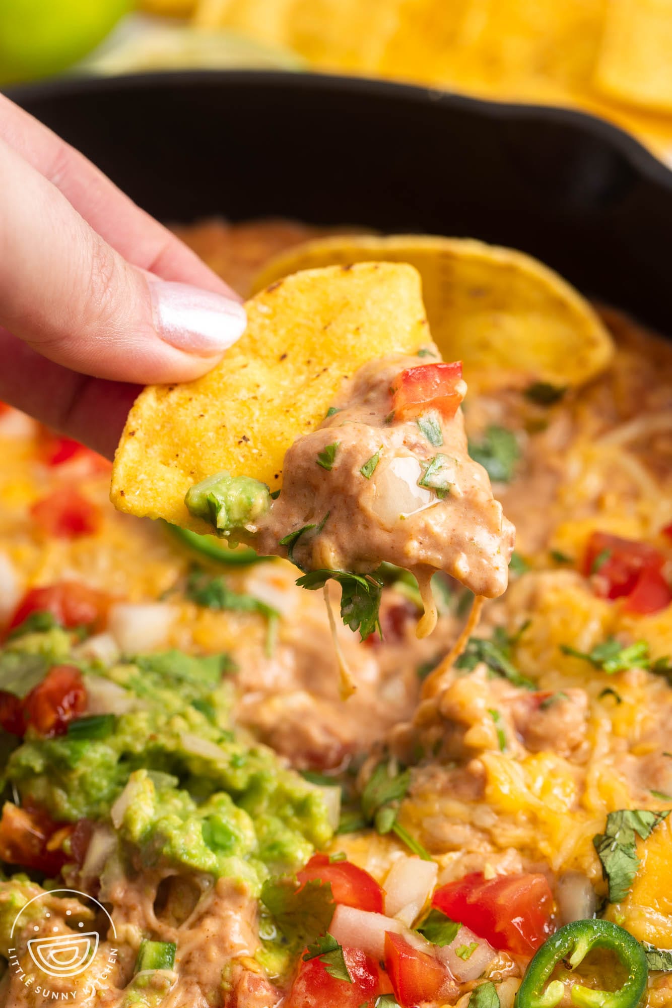 a hand holding a tortilla chip that has been dipped into cream cheese bean dip with pico de gallo and guac.