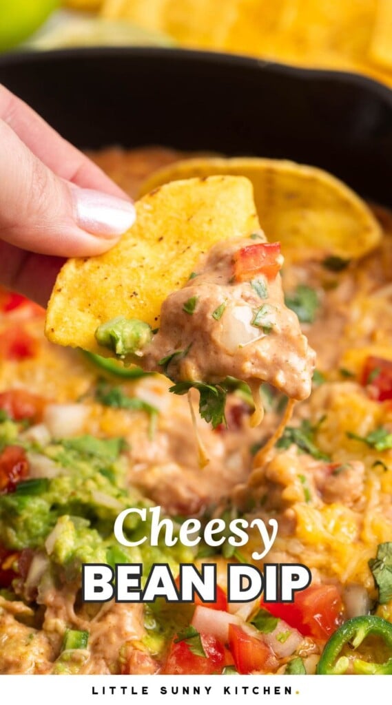 a hand holding a tortilla chip that has been dipped into cream cheese bean dip with pico de gallo and guac. Text overlay says "cheesy bean dip"