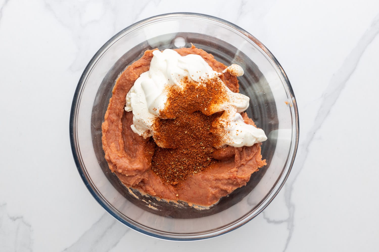 a glass mixing bowl holding refried beans, cream cheese, sour cream, and taco seasoning. 