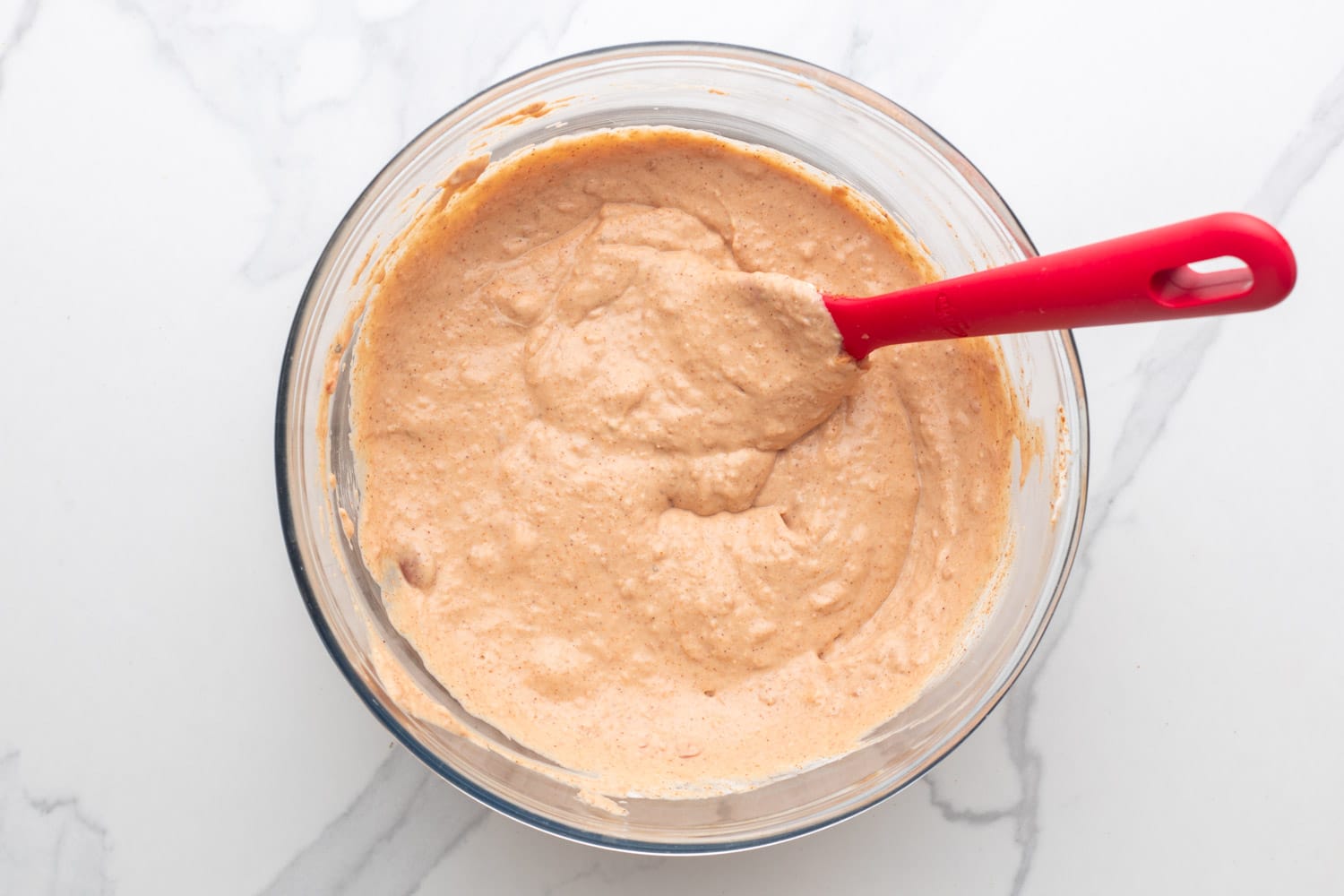 creamy bean dip mixture in a glass mixing bowl, stirred with a red spatula. 