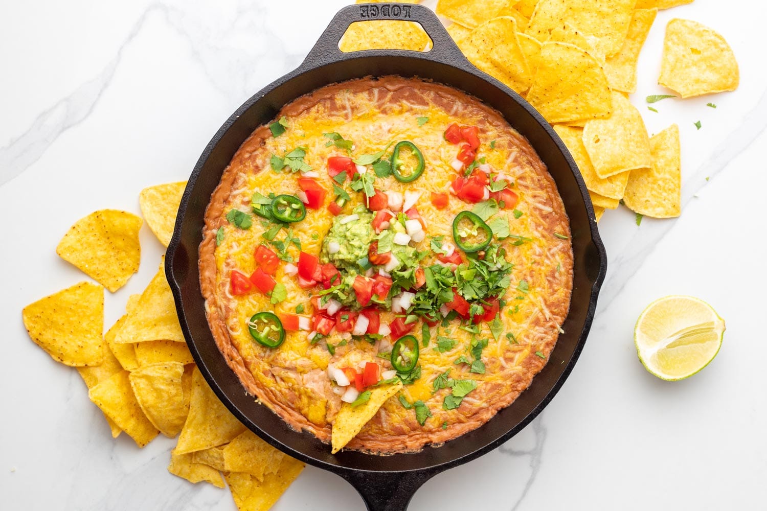 toppings added to cheesy bean dip in a lodge cast iron pan. around the pan are corn tortilla chips and half a lime.