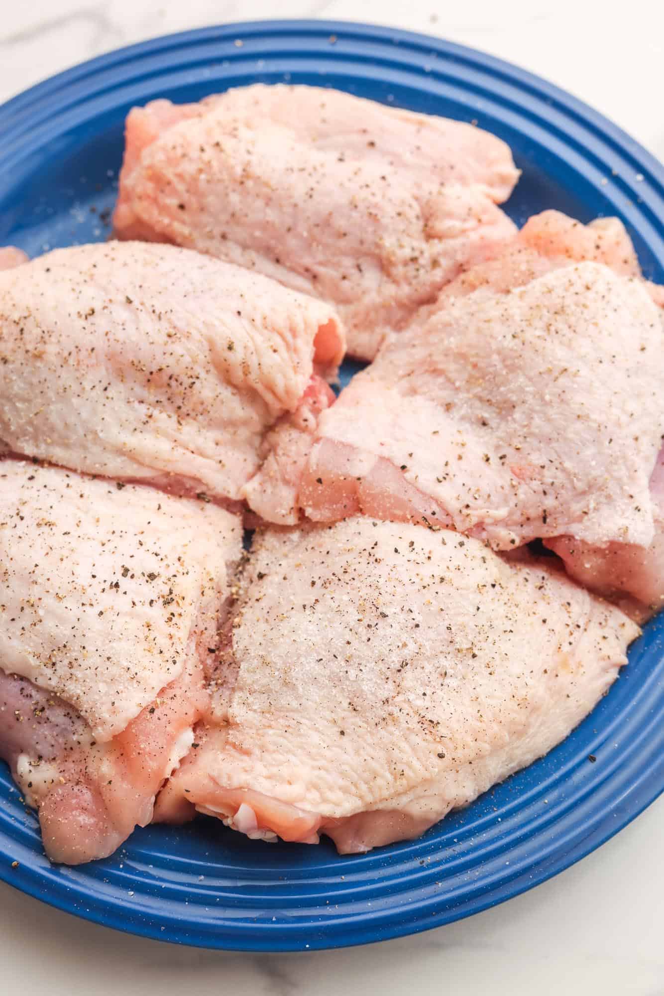 bone in skin on chicken thighs, seasoned with salt and pepper, on a blue plate.