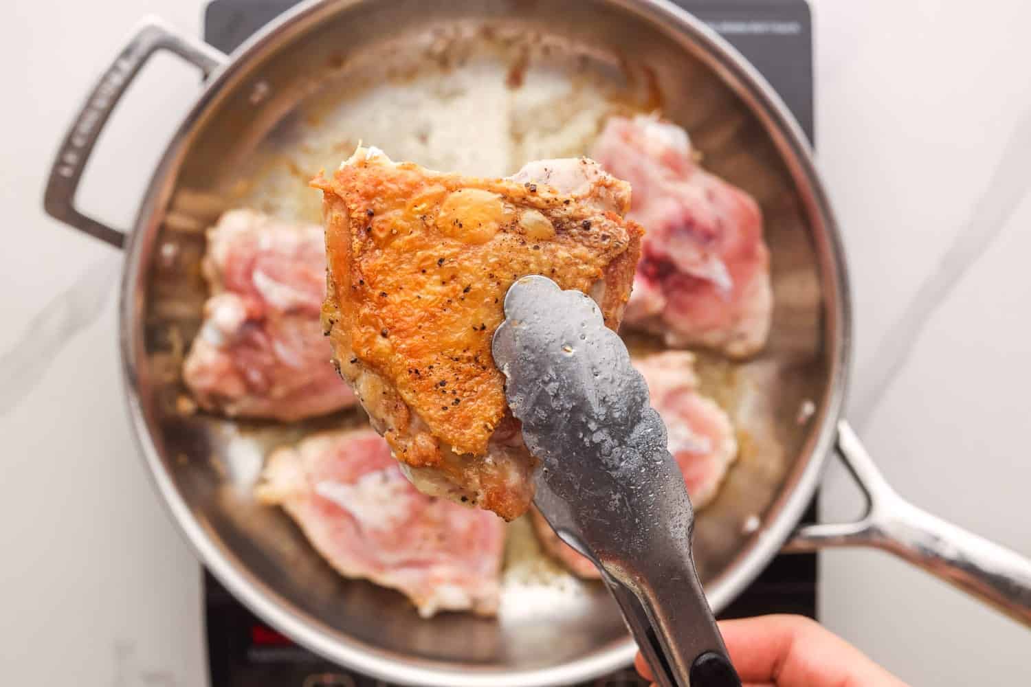raw chicken thighs in a stainless steel pan, cooking. One is picked up with tongs to show how the skin has been browned.