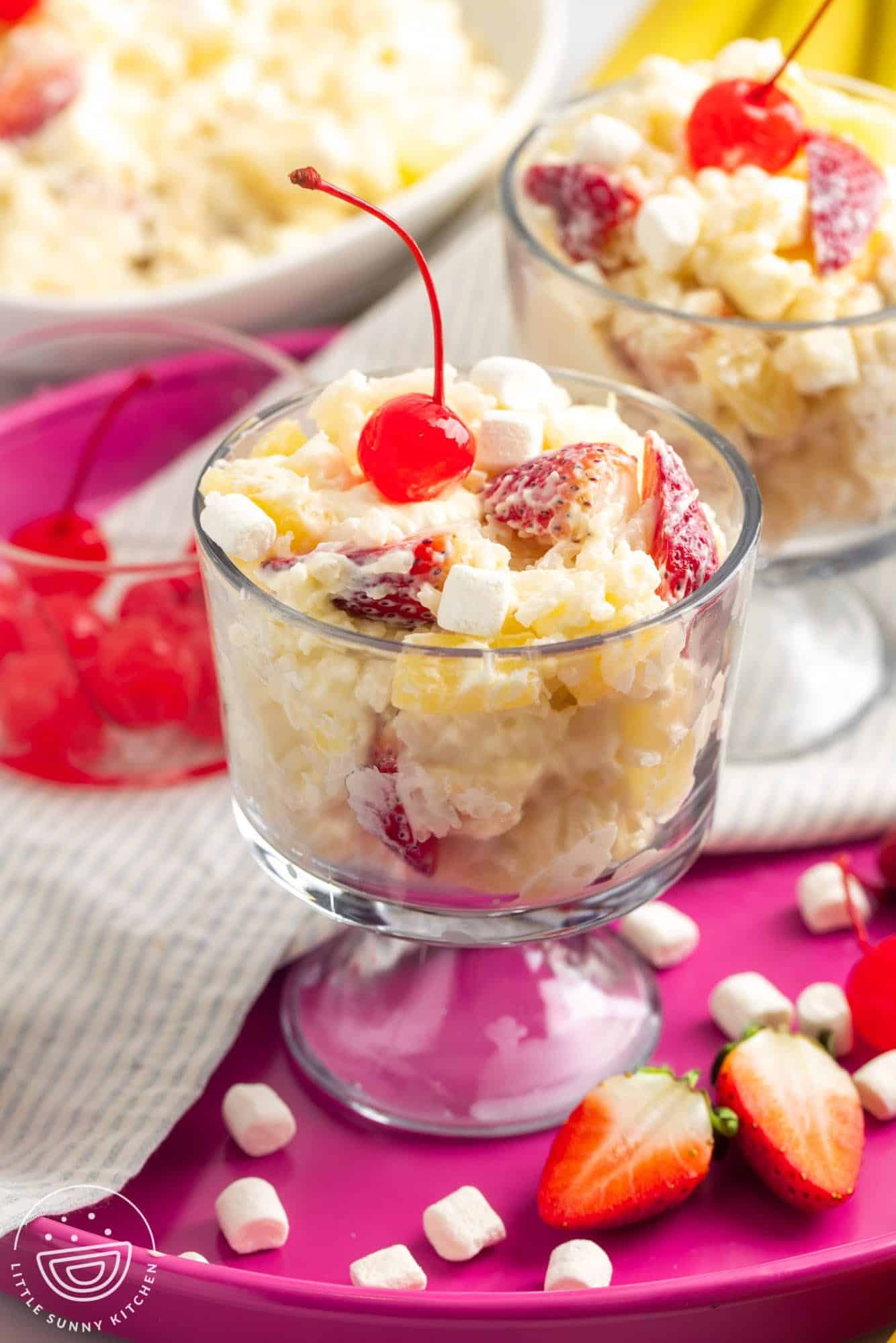 a footed glass dessert dish filled with creamy glorified rice with fruit and marshmallows. Topped with a maraschino cherry.