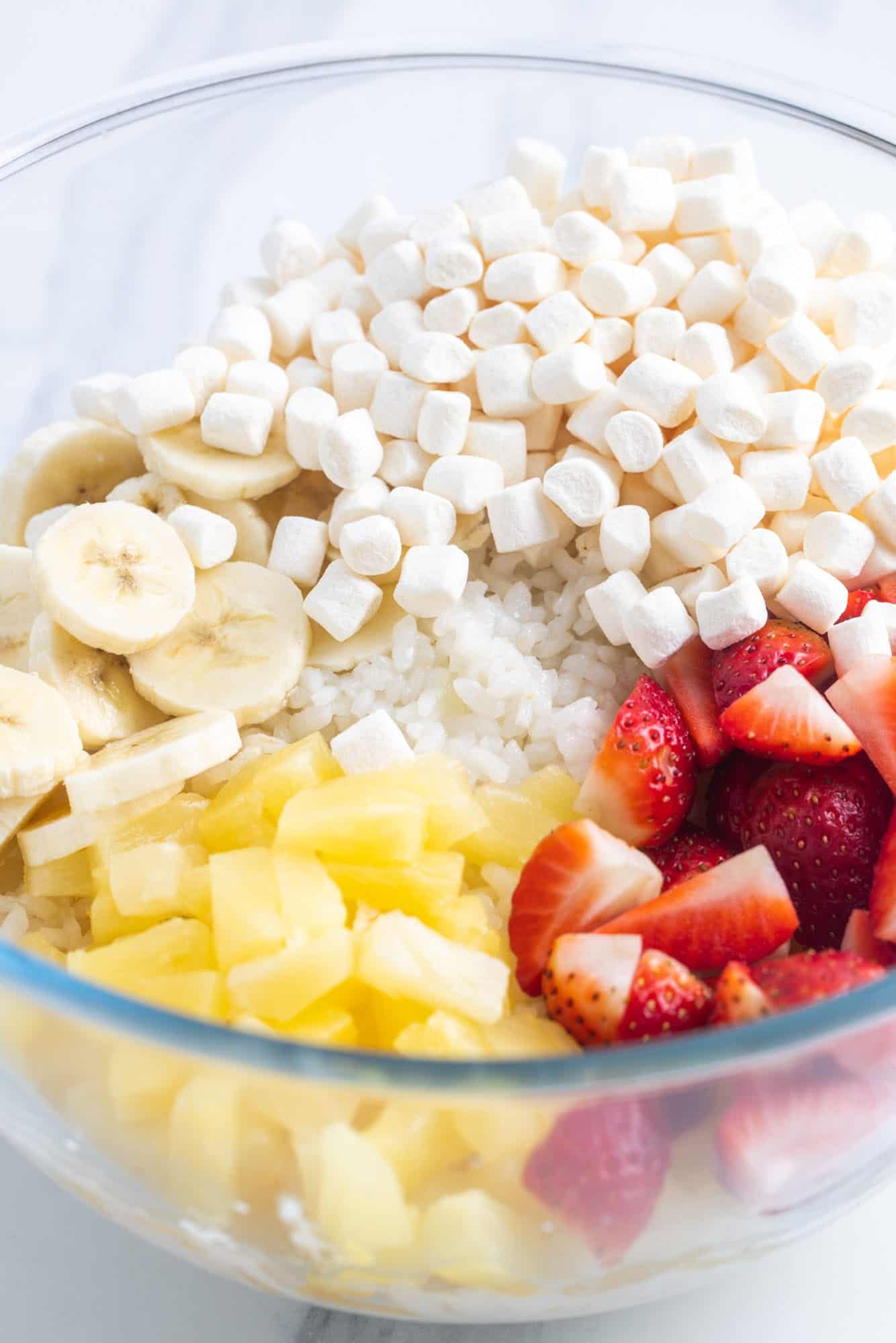 a glass bowl of cooked rice, topped with mini marshmallows, sliced strawberries, sliced bananas, and pineapple tidbits.