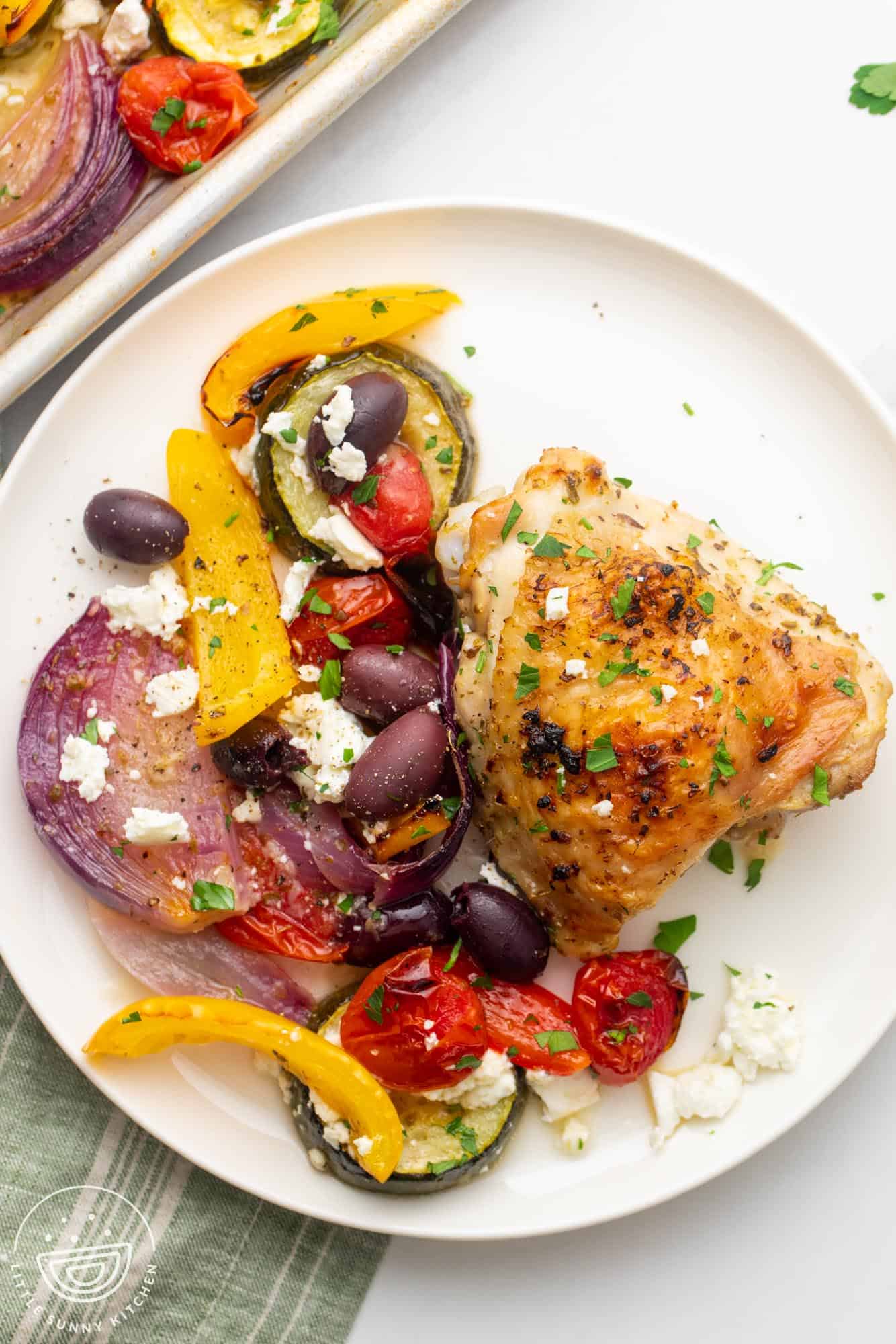 a white dinner plate with a greek marinated chicken thigh and roasted veggies with kalamata olives and feta cheese