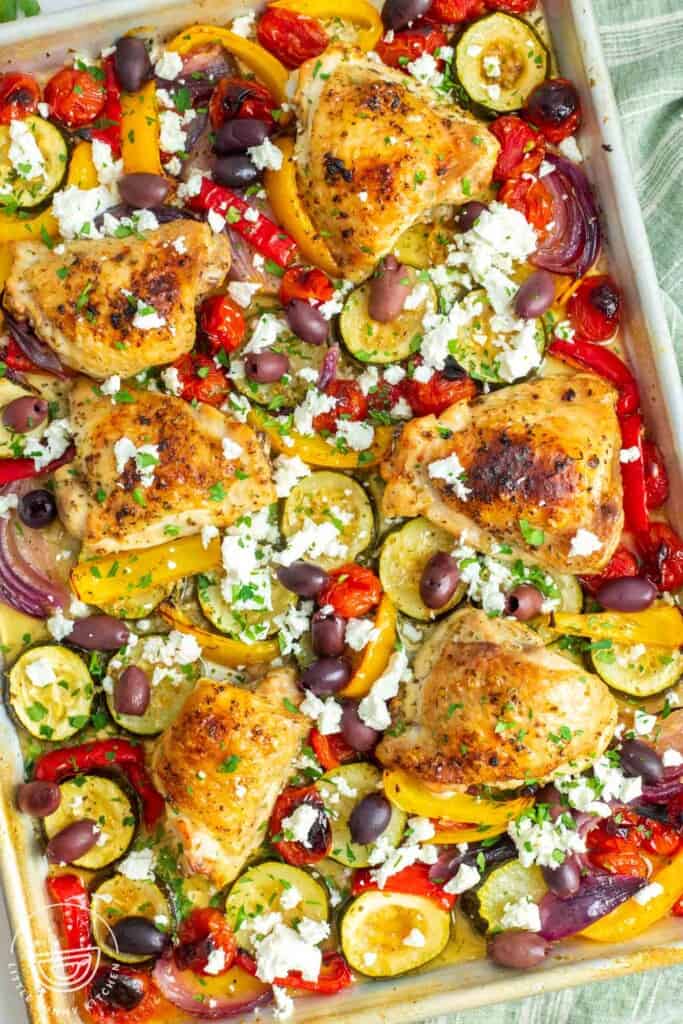bone in, skin on chicken thighs nestled on a sheet pan with tomatoes and peppers, kalamata olives, and feta cheese.