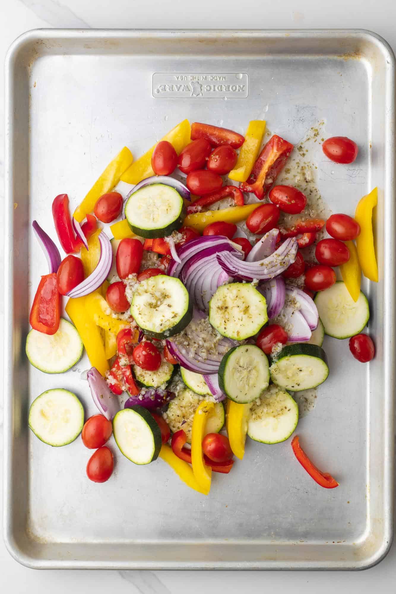 sliced zucchini, bell peppers, onions, and whole cherry tomatoes tossed with green marinade on a metal sheet pan. 