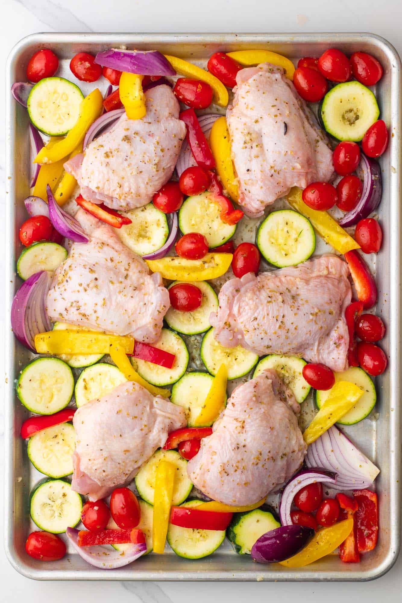 marinated chicken thighs on top of vegetables on a sheet pan. 
