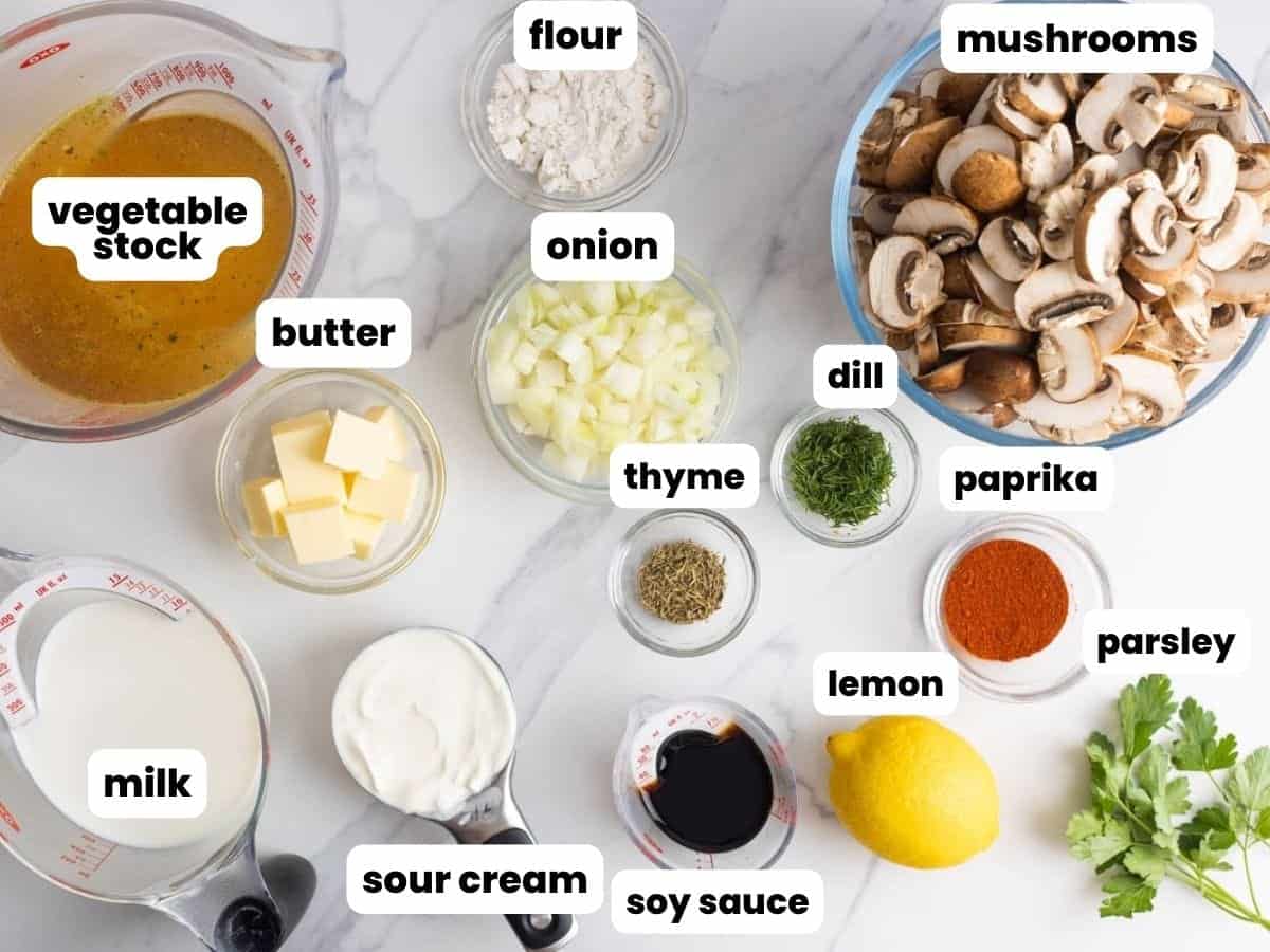 All of the ingredients for making homemade hungarian mushroom soup, measured into small bowls on a marble counter. There is a large bowl of sliced mushrooms, a pitcher of vegetable stock, and small bowls with seasonings, sour cream, butter, and milk.