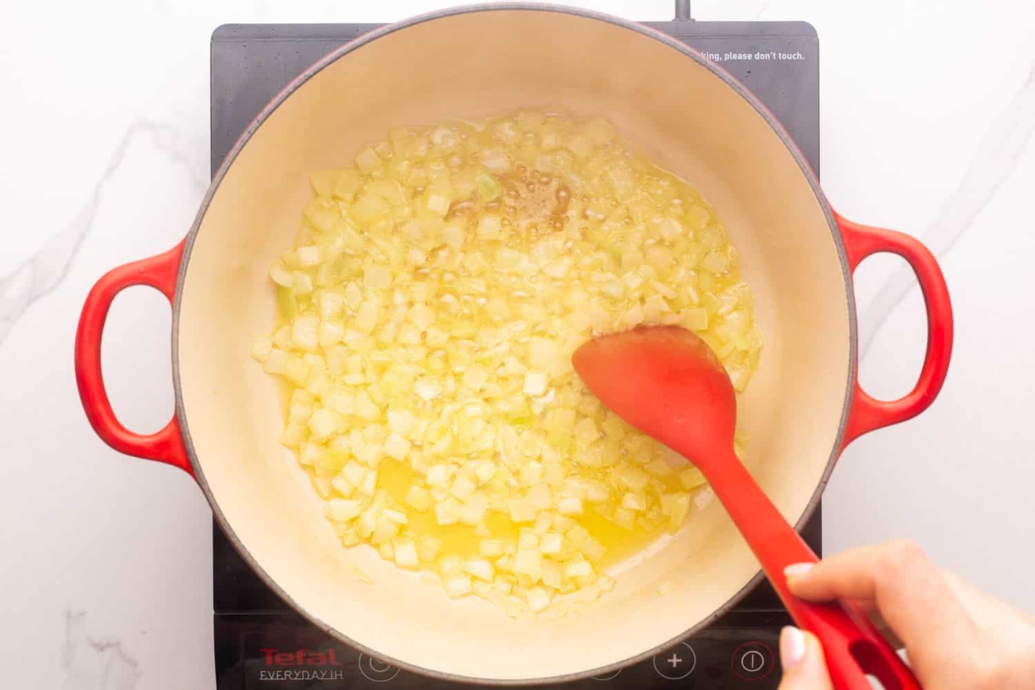 diced onions sauteed in butter in a ceramic coated cast iron dutch oven. viewed from above, the onions are being stirred with a red spoon.