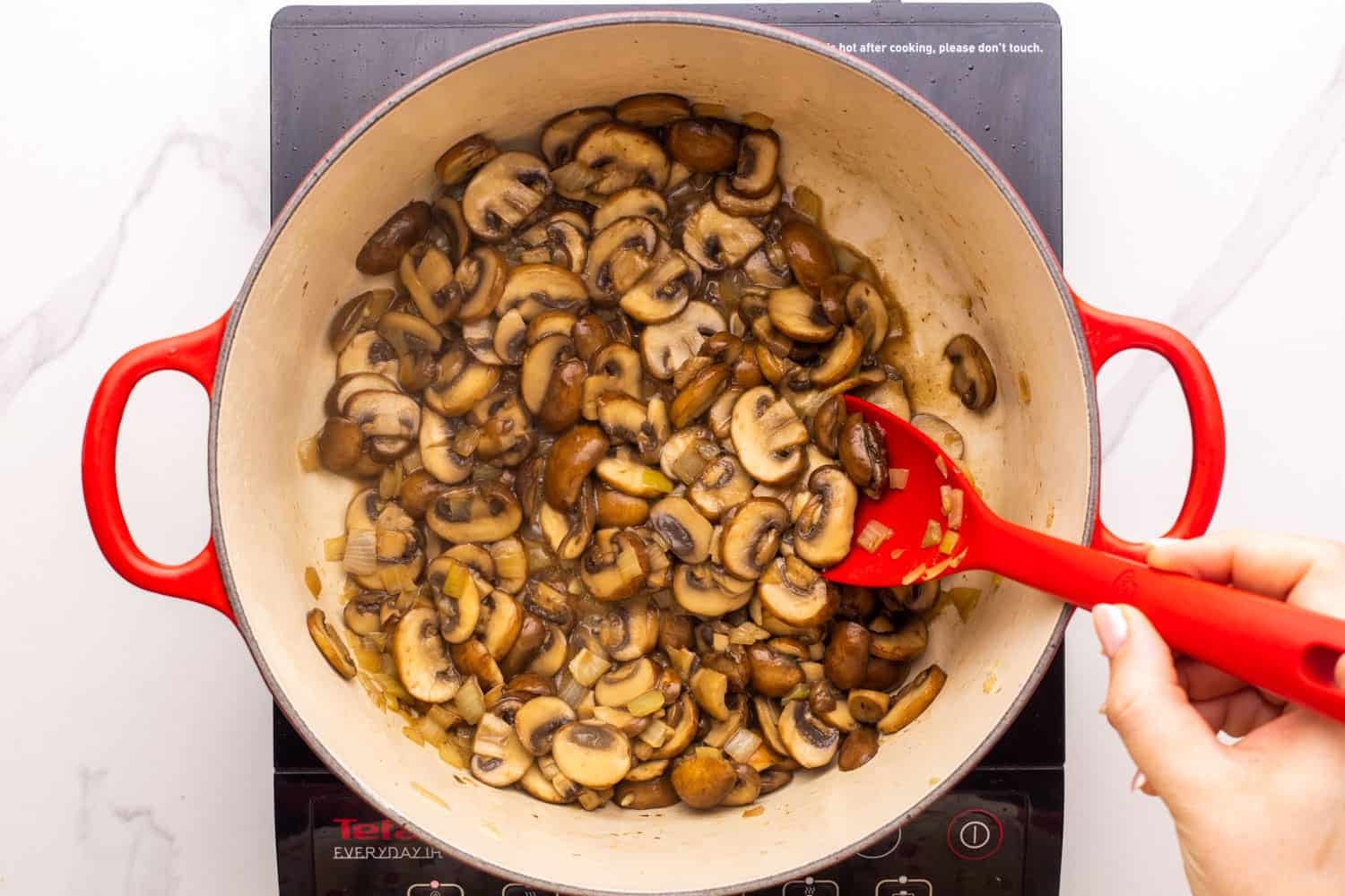 sliced cremini mushrooms cooking in a dutch oven. viewed from above, the mushrooms are browned and stirred by hand with a red spoon.