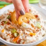 a bowl of creamy dip with cheese, bacon, green onions and almonds. A hand is dipping two ritz crackers into the bowl.