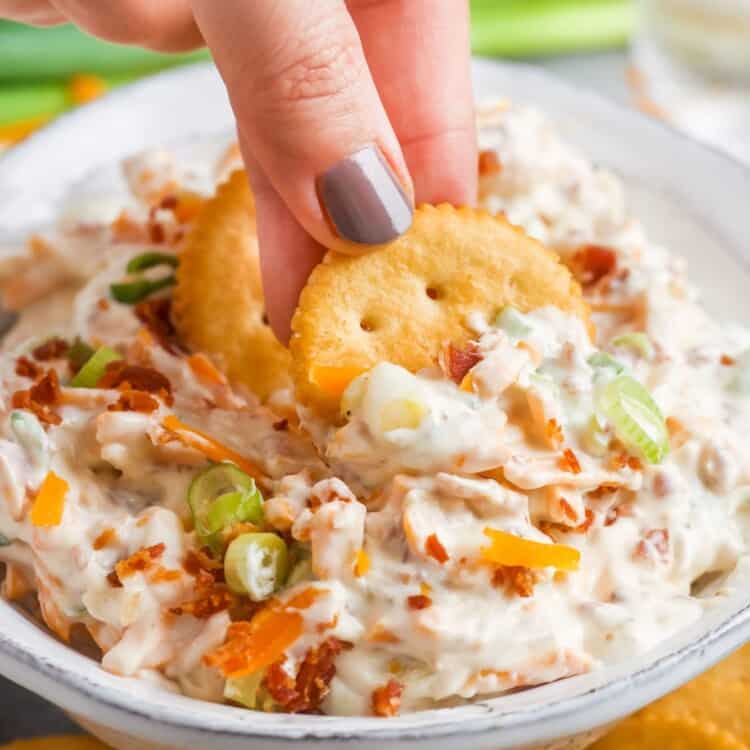 a bowl of creamy dip with cheese, bacon, green onions and almonds. A hand is dipping two ritz crackers into the bowl.