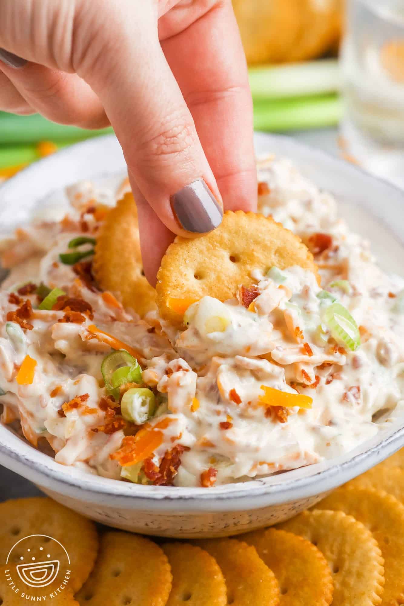 a bowl of creamy dip with cheese, bacon, green onions and almonds. A hand is dipping two ritz crackers into the bowl.