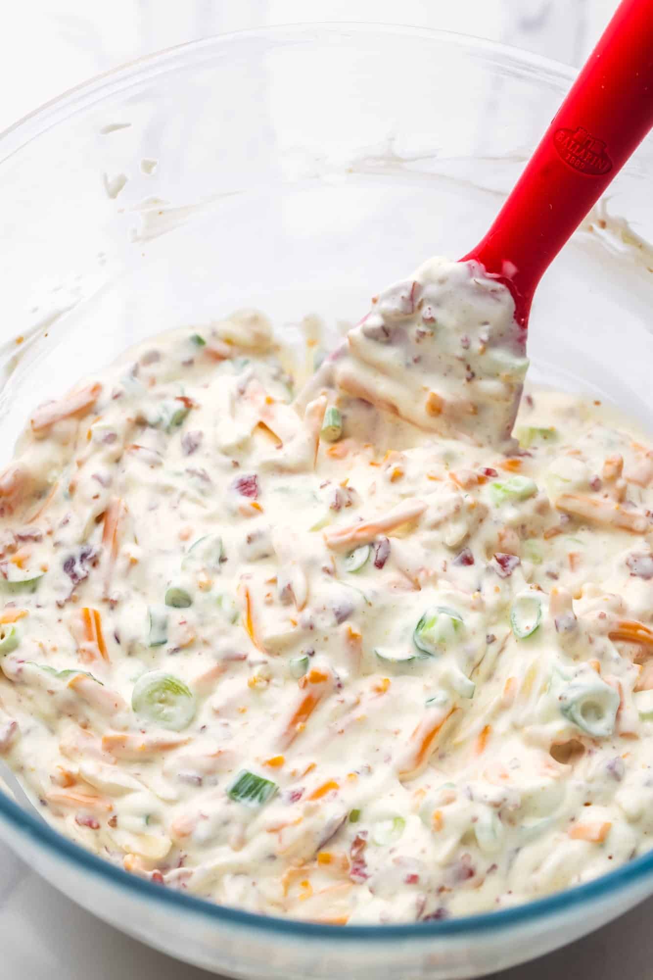 a glass mixing bowl of creamy million dollar dip with a red spatula in it. 