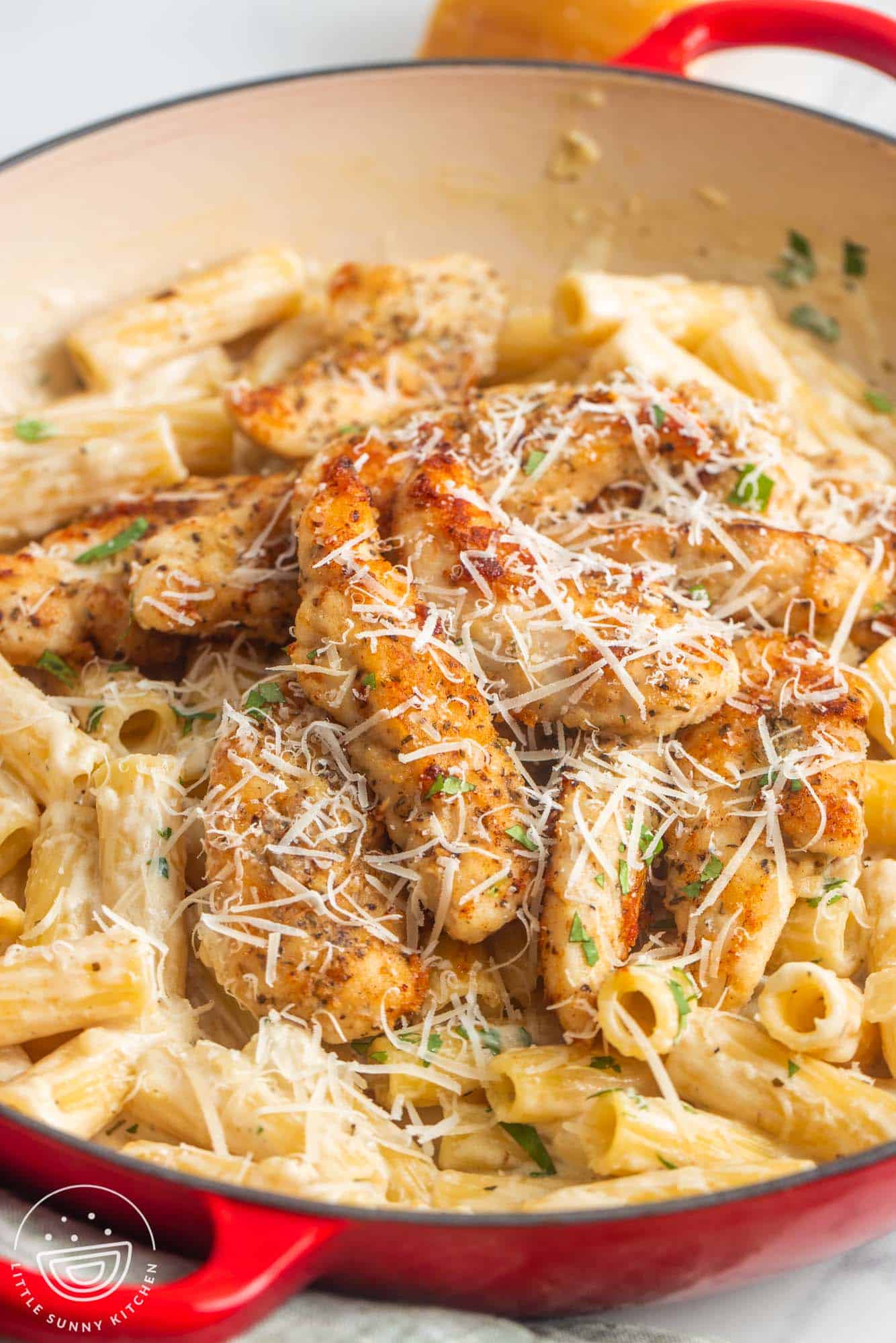 a red enameled skillet of parmesan chicken tenderloins over creamy ziti pasta. The dish is topped with fresh parsley and grated cheese.