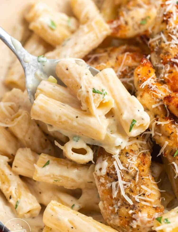a spoon holding creamy ziti pasta with parmesan cheese, picked up from a skillet filled with pasta and pan seared chicken tenders.