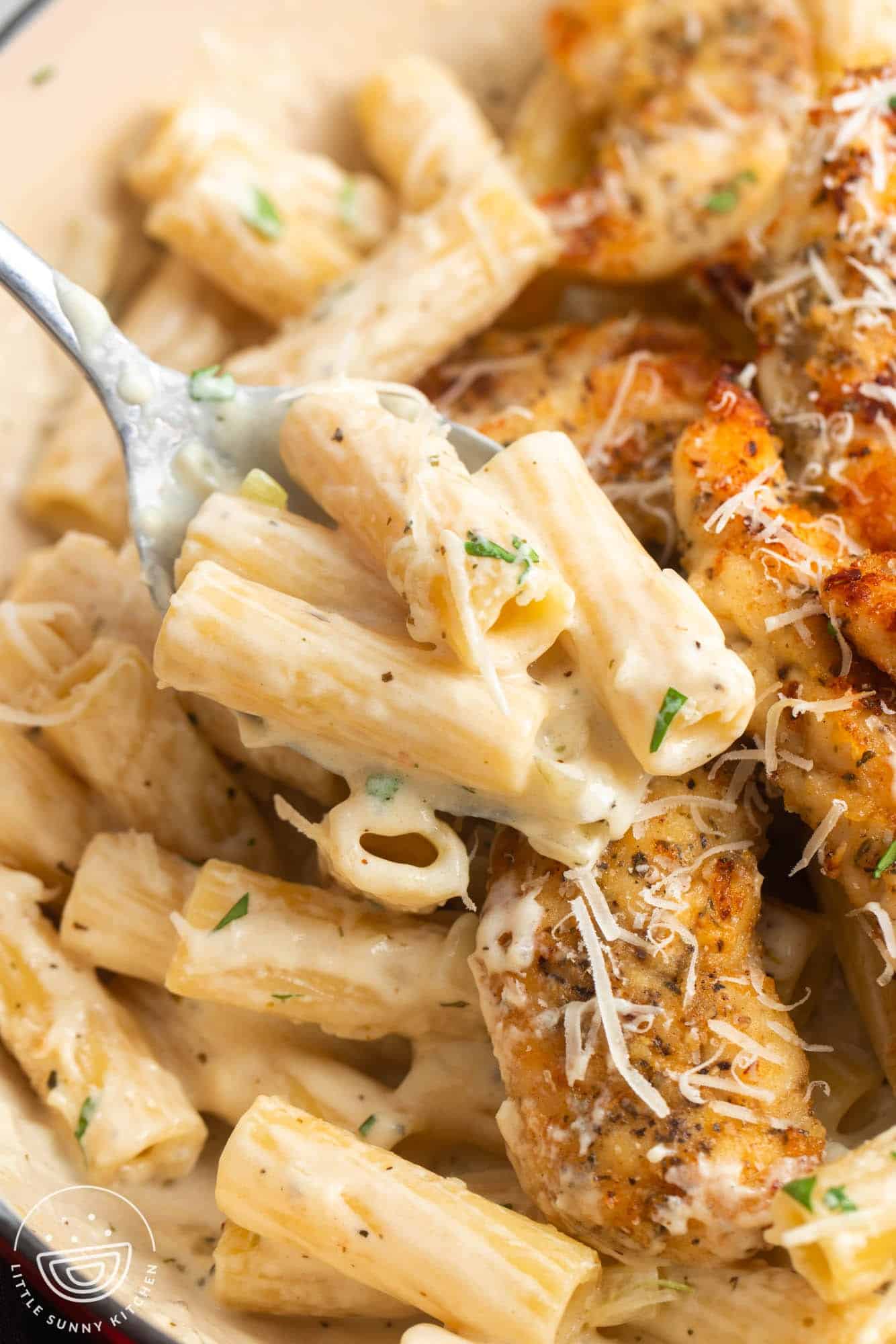 a spoon holding creamy ziti pasta with parmesan cheese, picked up from a skillet filled with pasta and pan seared chicken tenders.