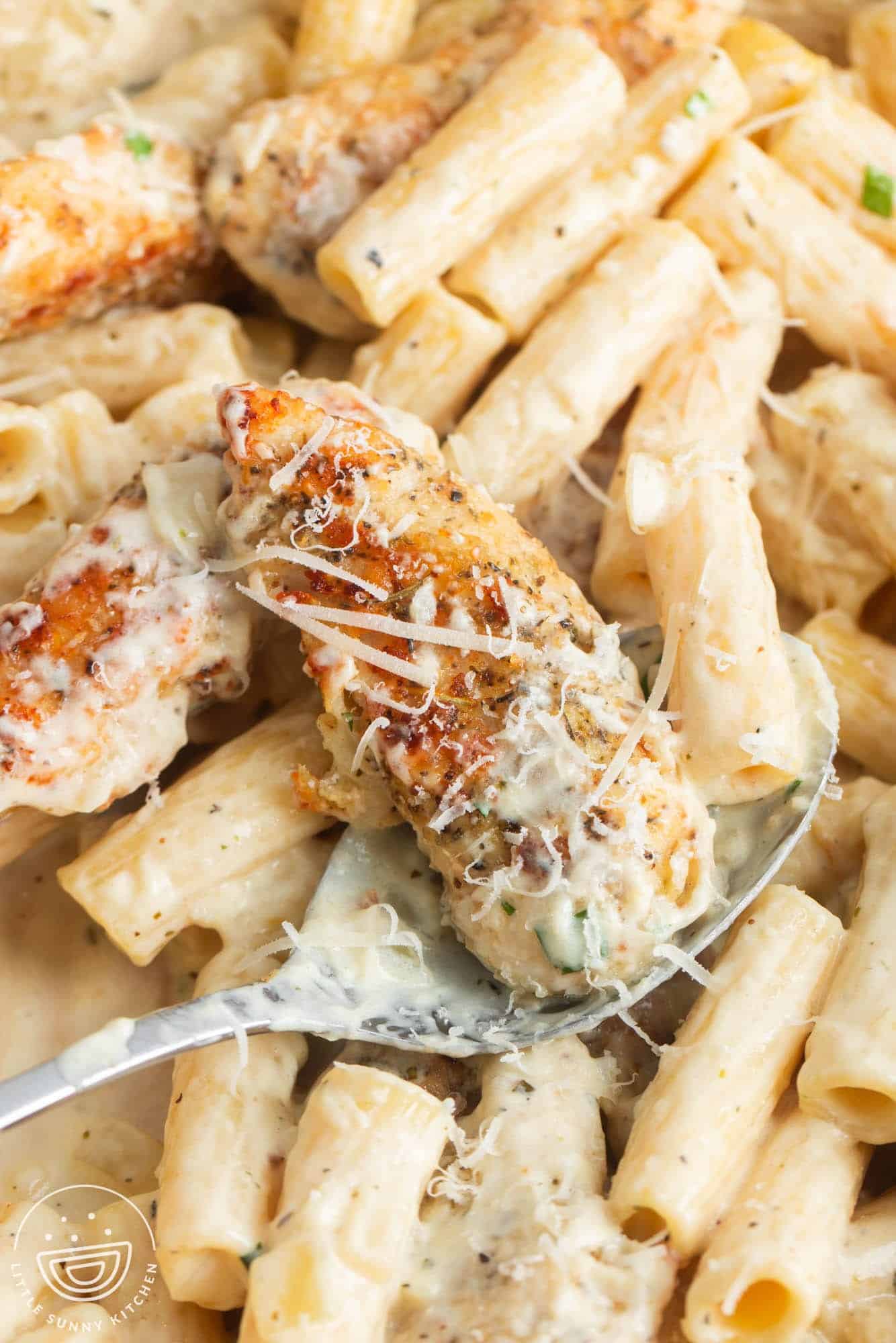 closeup of a pan of creamy parmesan chicken pasta. A spoon is holding up a piece of chicken that is topped with grated parmesan cheese.