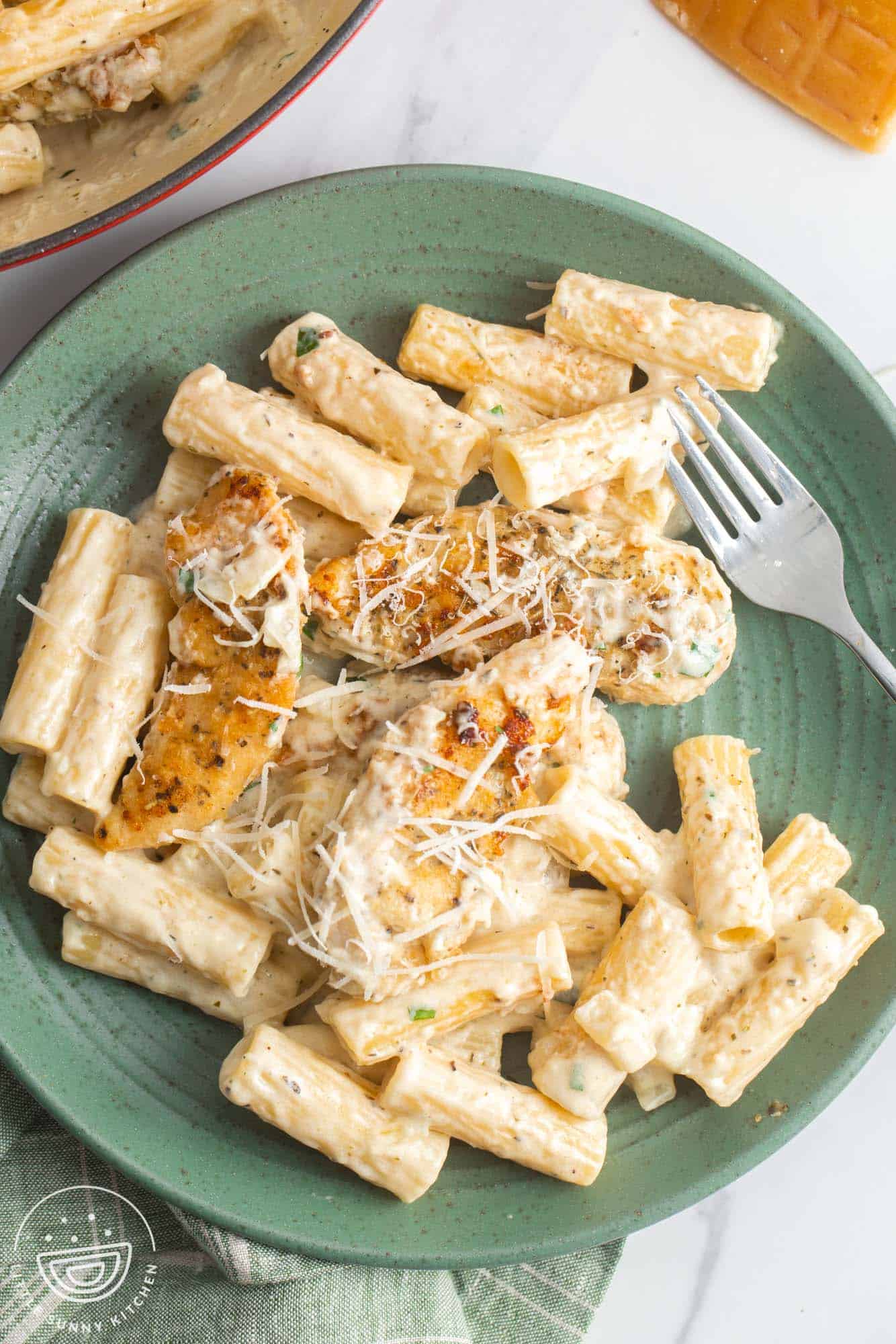 a green ceramic plate holding creamy parmesan tube pasta with three pieces of chicken, all topped with sauce and parmesan cheese.