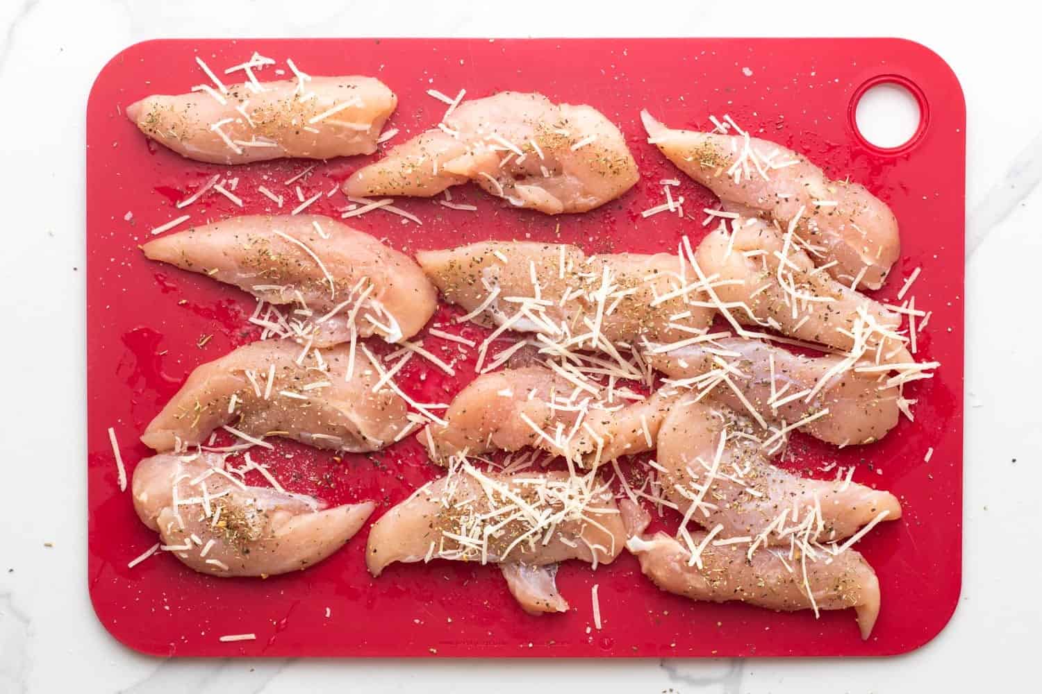 a red plastic cutting board holding 13 chicken tenderloins that have been seasoned with italian seasoning and parmesan cheese shreds.