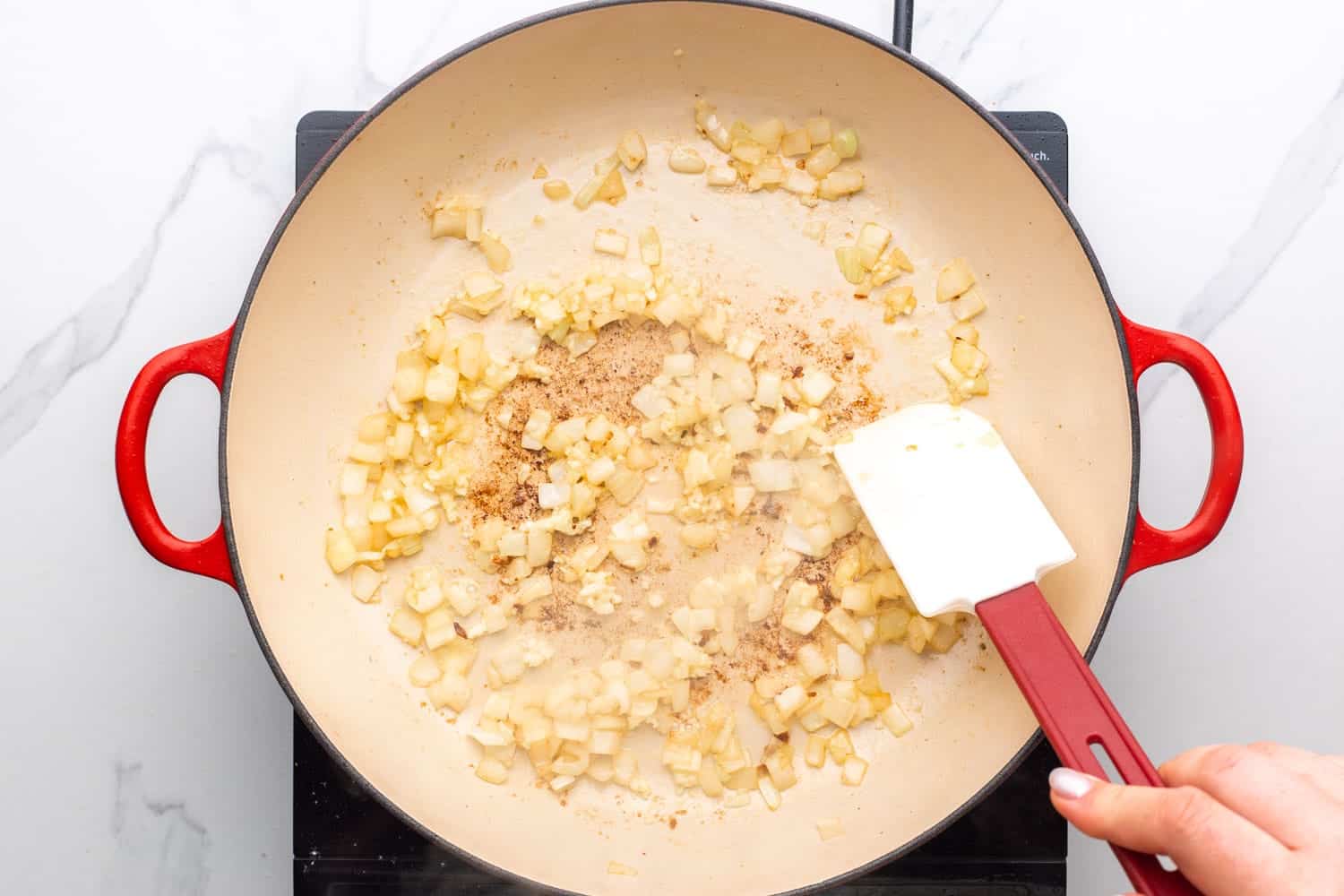overhead view of diced onion and garlic cooking in an enamel coated skillet, stirred with a red and white spatula.
