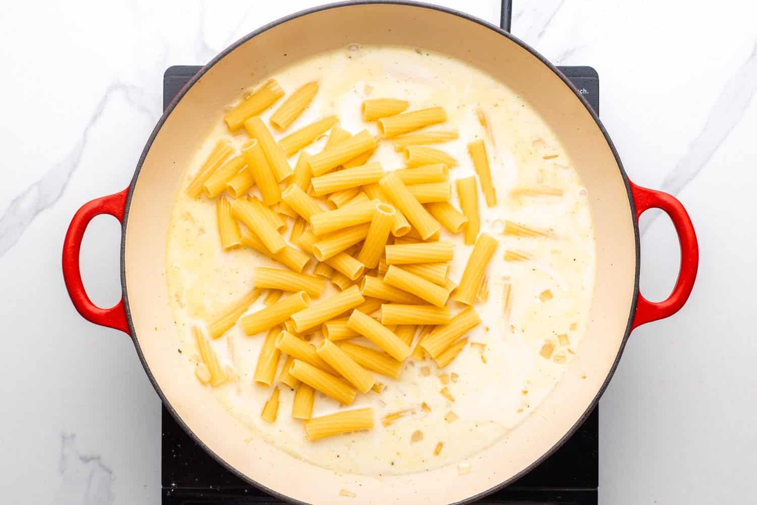 uncooked pasta added to a creamy parmesan sauce in a large skillet, viewed from above.