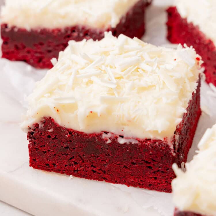 square red velvet brownies with frosting and shaved white chocolate sprinkles, arranged on a platter.