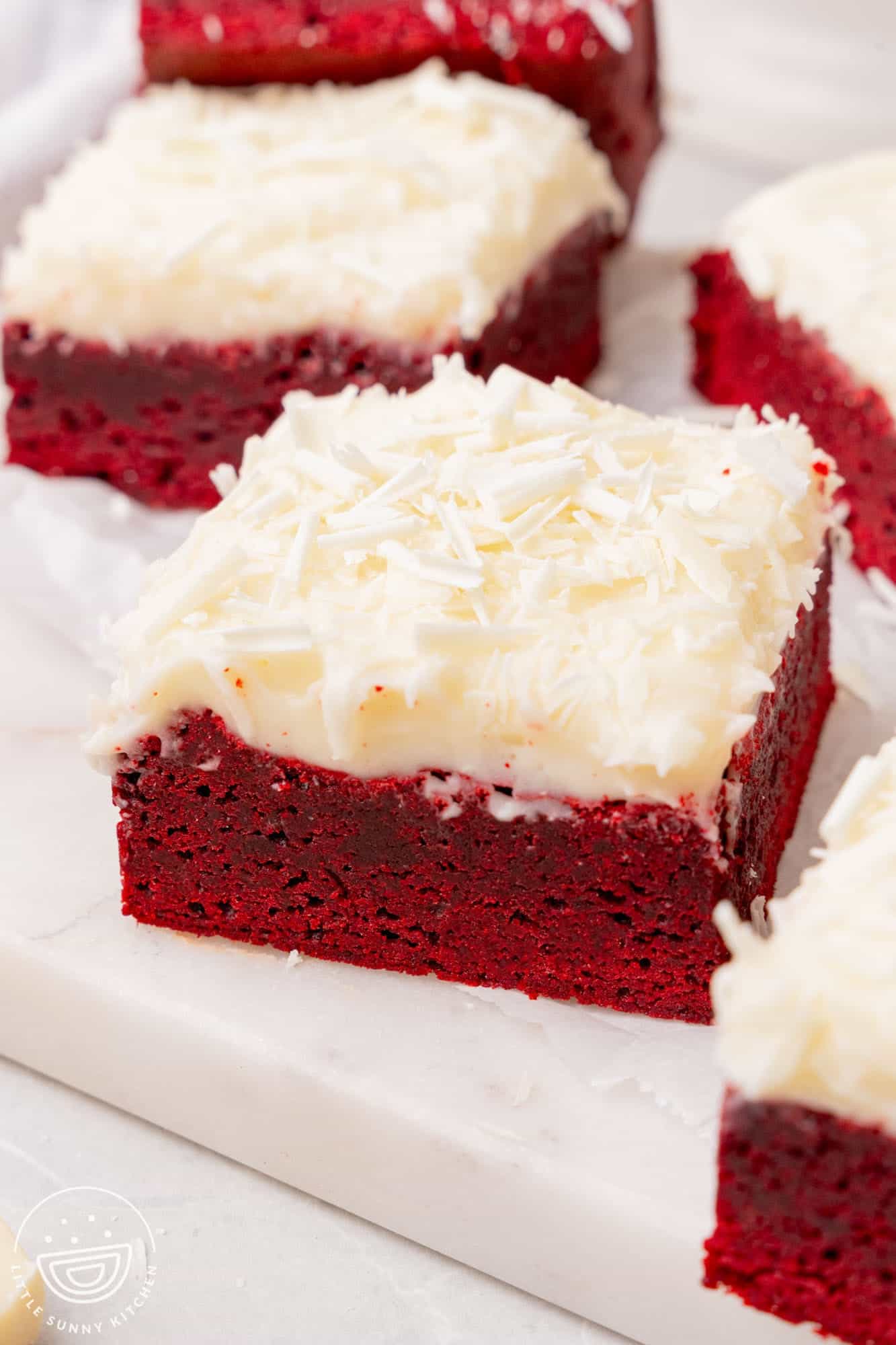 square red velvet brownies with frosting and shaved white chocolate sprinkles, arranged on a platter.