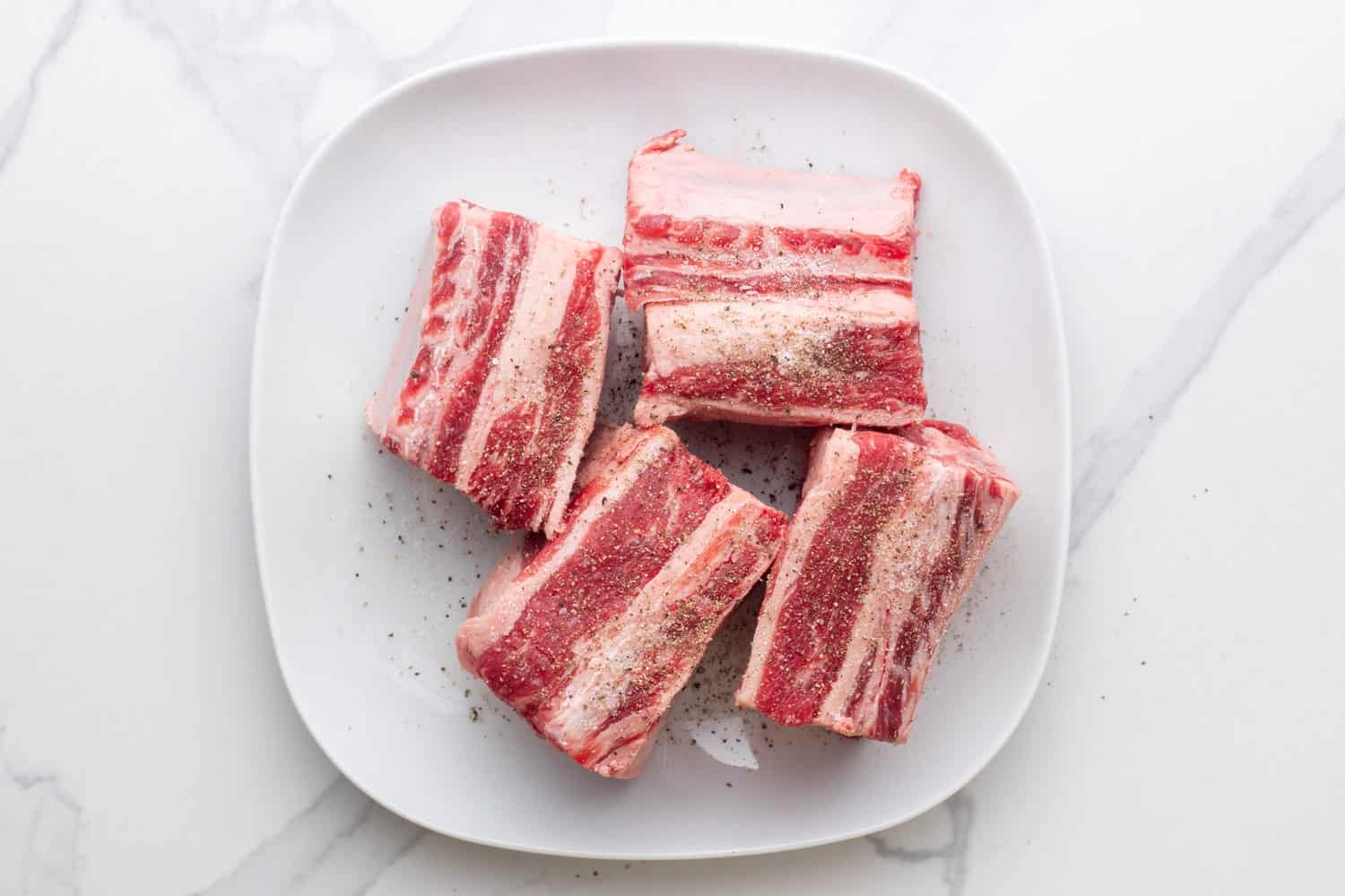 four pieces of beef short ribs on a square plate, seasoned with salt and pepper. 