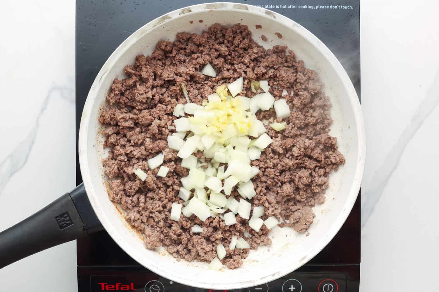 a white skillet of ground beef with chopped onion and minced garlic. 